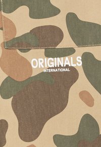 Tessuto beige con motivo mimetico in tonalità di verde, marrone e sabbia. Il testo "ORIGINALS INTERNATIONAL" è stampato in bianco.