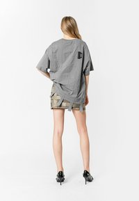 Camisa gris de gran tamaño con bolsillos de parche y detalles de lazo en la parte posterior, combinada con una falda beige en capas. La modelo lleva zapatos de tacón negros.