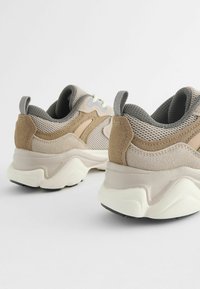 Beige og tan sneakers med mesh- og ruskindsmaterialer, teksturerede accenter og en kraftig hvid sål med sorte skridsikre. 