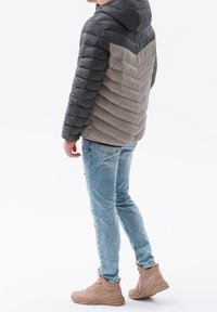 Muž v dvoubarevném černobílém péřovém kabátu, světlých modrých skinny džínách a béžových vysokých teniskách stojící na bílém pozadí.
