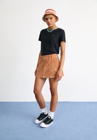 Zwarte korte mouwen t-shirt, tan corduroy rok met voorzakken, zwarte sneakers en een meerkleurige gestreepte bucket hoed, afgebeeld op een lichte achtergrond.