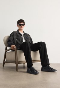 Chaqueta de mezclilla negra, camiseta blanca, pantalones negros y zapatillas negras brillantes. Sentado en una silla de color claro contra un fondo sencillo.