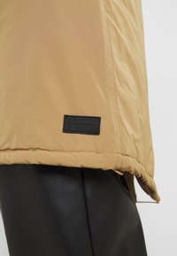 Ourlet d'une veste beige avec une étiquette noire "Scotch & Soda" portée par-dessus un pantalon en cuir noir.