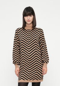 Kvinde iført en langærmet kjole med beige og sort chevron-stribede mønstre, med puffede ærmer og en høj halsudskæring, stående mod en ensfarvet baggrund.