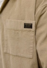Tan corduroy shirt met een voorzak, voorzien van een zwart label met gouden tekst. De stof heeft verticale ribbels en een zachte textuur.