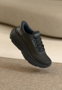 Μαύρο αθλητικό παπούτσι με στρογγυλή μύτη, διχτυωτό επάνω μέρος και παχύ πάτο με αυλακώσεις. Επωνυμία στο πλάι σε γκρί, ορατή υφή από υφασμάτινο υλικό.