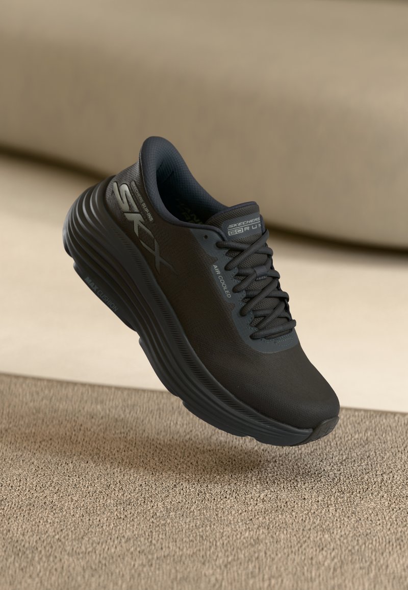 Shoe de corrida preto com bico arredondado, parte superior em malha e sola grossa com ranhuras. Marca na lateral em cinza, detalhe em tecido texturizado visível.