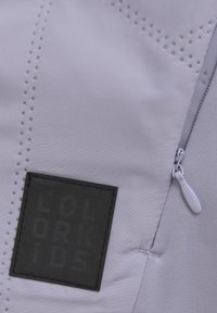 Grått tyg med subtila perforeringar och en svart patch med texten "COLOR KIDS". Dragkedjan har en vit dragare. Strukturerad ytdetalj synlig.