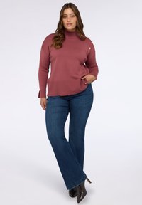 Staubrosafarbener Rollkragenpullover mit seitlichen Knöpfen, kombiniert mit dunkelblauen Schlagjeans und spitzen schwarzen Heels. Strukturierter Strickstoff.