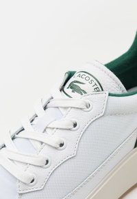 Beli superge z teksturirano usnje in mrežasto zasnovo, zelenimi detajli, belimi vezalkami in okroglo Lacoste logotipom na jezičku.