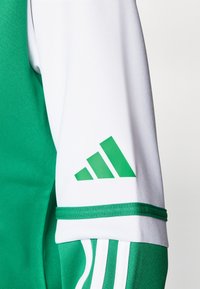 Grøn og hvid atletiktrøje med struktureret stof, tre hvide striber og et grønt trekantet logodetalje på ærmet.