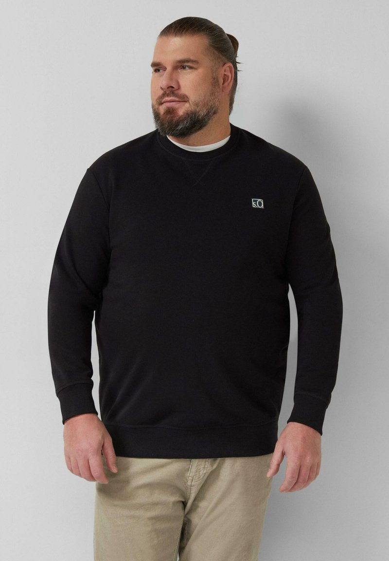 Bärtiger Mann mit zurückgebundenem Haar, trägt ein schwarzes Sweatshirt mit langen Ärmeln und beige Hosen, steht vor einem einfarbigen hellgrauen Hintergrund.