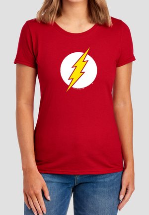 Frau trägt ein rotes T-Shirt mit einem gelben Blitz in einem weißen Kreis vorne, kombiniert mit blauen Jeans.