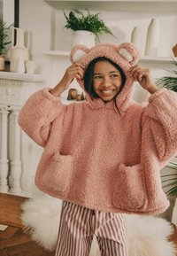 Rosa flauschiger Poncho mit Kapuze und Bärenohren, weicher Textur, zwei Fronttaschen und weiten Ärmeln. Kombiniert mit rosa und weißen gestreiften Hosen.