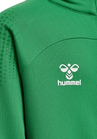 Grøn atletisk hættetrøje lavet af tekstureret stof. Har et hvidt logo og et mønstret skulderdesign, der fremhæver åndbarheden.