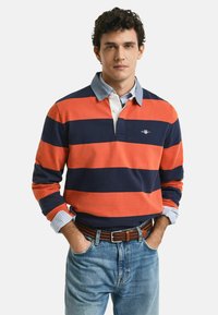 Camisa de polo de manga larga en rayas naranja y azul marino, con un cuello blanco y puños a cuadros azules. Combinada con jeans azul claro y un cinturón marrón.
