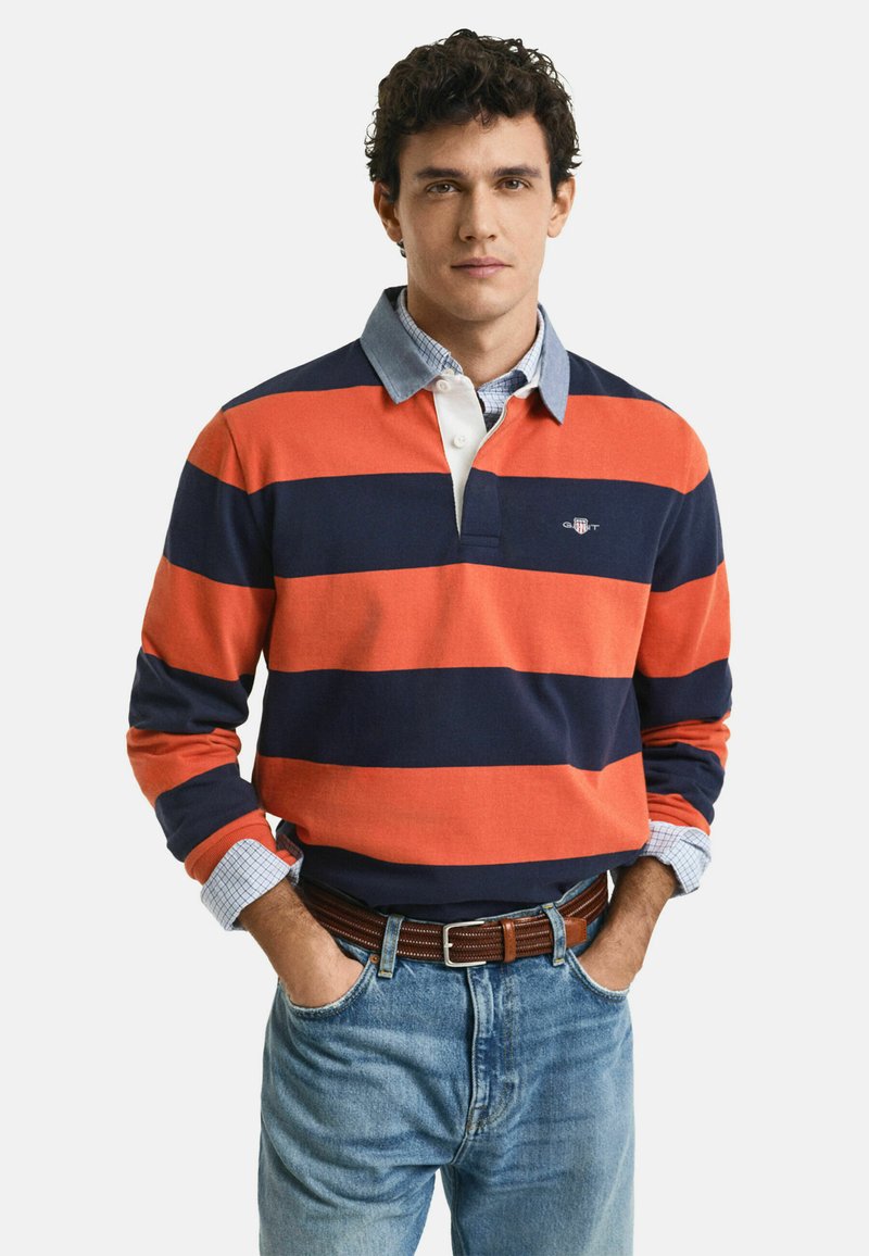 Camisa de polo de manga larga en rayas naranja y azul marino, con un cuello blanco y puños a cuadros azules. Combinada con jeans azul claro y un cinturón marrón.