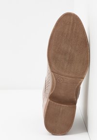 Semelle d'une chaussure beige avec un motif ondulé et la taille 37, placée à la verticale contre un mur blanc en angle.