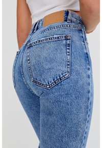 Jean en denim bleu taille haute avec un délavage, coutures doubles et un patch en cuir beige à l'arrière de la taille. Comprend deux poches arrière.