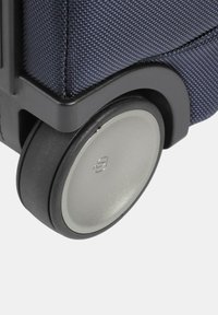Angolo di una valigia in tessuto blu navy con bordo in plastica nera e una ruota argentata. La ruota ha una superficie liscia e una marca discreta.