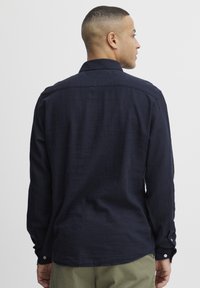 Camisa de manga larga azul marino en tejido ligero, con cuello clásico, puños abotonados y costura trasera recta.