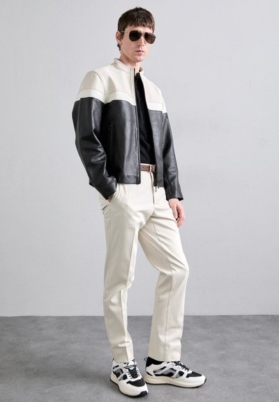 Black and cream leather jacket with white stripe, black shirt, beige trousers, sunglasses, and black-and-white sneakers. Textured materials featured.
