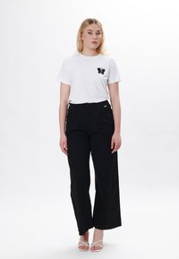 Hvid t-shirt med sort sommerfuglsbroderi på brystet, parret med høj talje sorte brede bukser og stribede hvide sandaler.