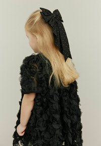 Crna cvjetna haljina od prozirnog materijala s 3D detaljima ruže. Velika crna mašna s točkastim uzorkom zavezana je u kosi, dodajući dekorativni naglasak.