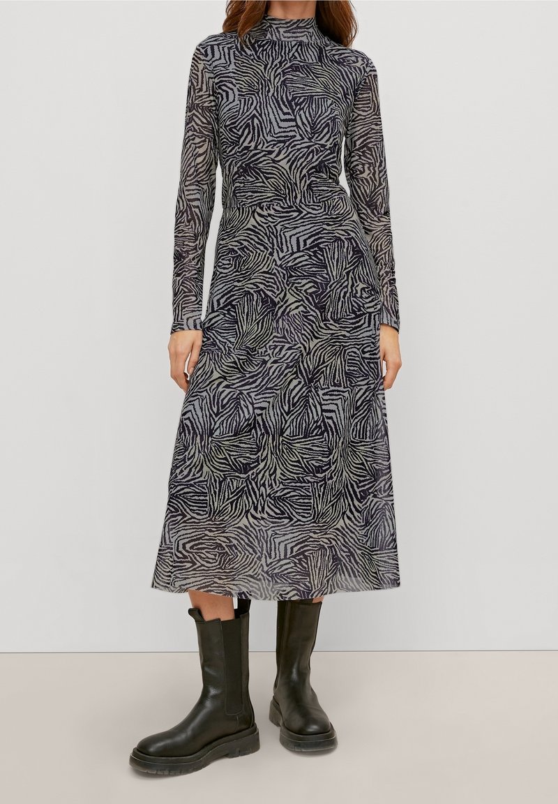 Vestido de manga larga en tela negra transparente con rayas abstractas en gris y púrpura imitando cebra. Presenta un escote alto y cae hasta la mitad de la pantorrilla. Llevado con botas negras.
