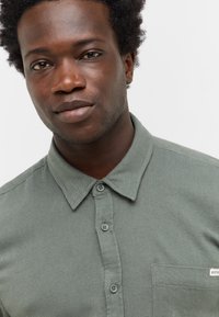 Bawełniana koszula zapinana na guziki w stonowanym zielonym kolorze, z kołnierzykiem, kieszonką z przodu i teksturowanym materiałem. Posiada szare guziki oraz subtelną metkę marki.
