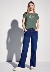 Olivově zelené tričko s grafickým textem, které je kombinováno s tmavě modrými kalhotami s širokými nohavicemi. Bílé tenisky dotvářejí outfit a mají hladkou texturu.