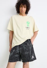 T-shirt oversize color crema con grafica di un cactus verde e testo, abbinata a pantaloni corti neri a fantasia con bordi bianchi e logo Adidas.