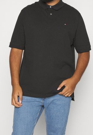 Mężczyzna w czarnej koszulce polo z krótkim rękawem i małym logo na piersi oraz jasnoniebieskich dżinsach na białym tle.