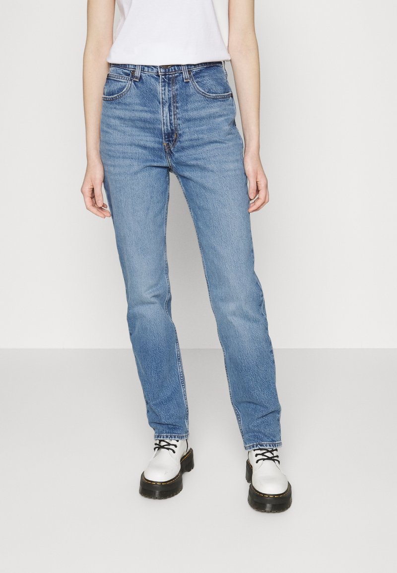 Personne portant un jean bleu clair droit, un haut blanc et des bottines blanches à lacets avec semelles noires, debout devant un fond uni.