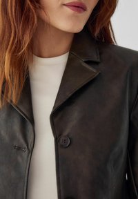 Barna bőrdzseki kis gallérral, két gombbal és sima textúrával, fehér felsőre rétegezve. Közelkép a nyakkivágásról és a szövet részleteiről.