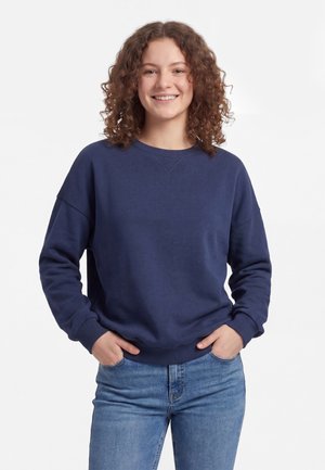 Ung person med krøllet hår iført en mørkeblå sweatshirt og blå jeans, smilende med hænderne i lommerne mod en ensfarvet hvid baggrund.