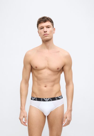 Μυώδης άνδρας που στέκεται μπροστά σε απλό φόντο, φορώντας λευκά εσώρουχα με μαύρη ζώνη μέσης που φέρει λευκά λογότυπα και γράμματα.