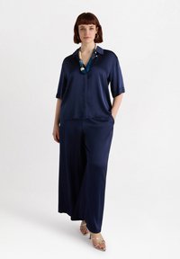 Completo in raso blu navy con maniche corte, vestibilità rilassata, pantaloni a gamba larga e una collana blu e bianca a più strati. Scarpe con tacco argentate.