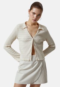 Camicia a maniche lunghe beige testurizzate con colletto e leggero arricciature, abbinata a una gonna avvolgente beige chiaro.