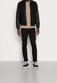 Svart bomberjacka med läderärmar, beige tröja, svarta jeans och svarta sneakers med vita sulor och snören, stående på en ljusgrå yta.