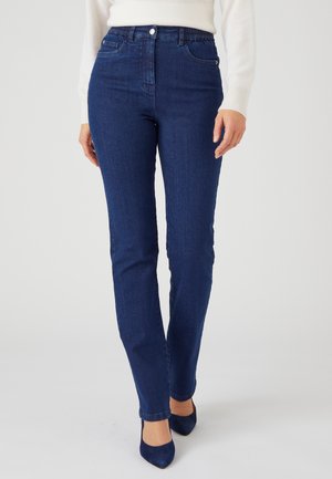 Femme portant un jean slim bleu foncé, un haut blanc à manches longues et des talons pointus bleu foncé, debout devant un fond uni.