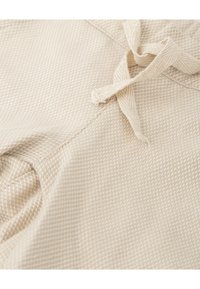 Tejido de color beige claro con textura que presenta un patrón tejido, con un detalle de cinta atada. Vista en primer plano que muestra la superficie y los detalles de la costura.