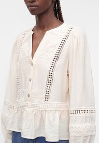 Femme portant un chemisier crème avec des panneaux en dentelle, des boutons sur le devant, des manches longues bouffantes et un bas évasé froncé.