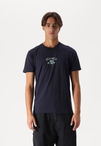 Marineblaue Baumwoll-T-Shirt mit kurzen Ärmeln, das ein zentrales grünes und weißes "STANCE"-Logo aufweist. Glatte Textur, Rundhalsausschnitt.