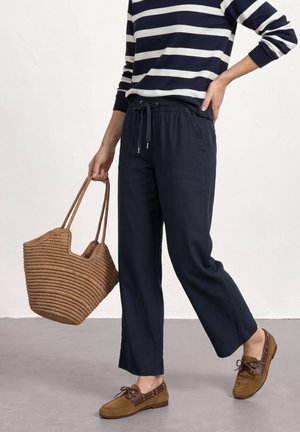 Personne portant un pantalon bleu marine à cordon et une chemise rayée à manches longues, tenant un grand sac fourre-tout marron tressé, portant des mocassins en cuir marron.
