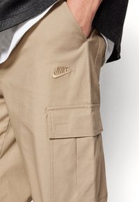 Béžové cargo kalhoty z texturované tkaniny, s výrazným logem Nike, přední kapsou a velkou postranní kapsou na nášivku.