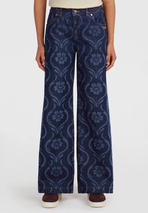Donkerblauwe wijde jeans met lichte bloem- en spiraalpatronen, voorzakken en bruine schoenen zichtbaar aan de onderkant.