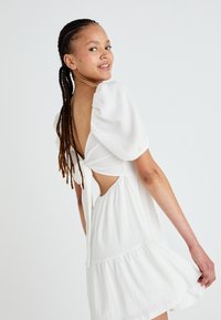 Λευκό κοντό φόρεμα με φουσκωτά μανίκια και ανοιχτή πλάτη, διαθέτει λεπτομέρεια με κομμένο κόμπο. Υφασμάτινη υφή με φούστα πολλαπλών των επιπέδων και ελαφριά ροή.