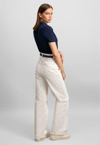 Marineblå kortermet polo-skjorte, hvite høyvidle bukser, flettet belte, lyse sneakers, minimalistisk bakgrunn.