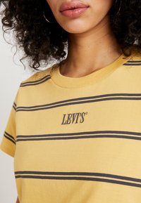 Žluté pruhované tričko vyrobené z bavlny, s horizontálními tmavě modrými pruhy a malým vyšívaným logem "LEVI'S" na hrudi. Kulatý výstřih.